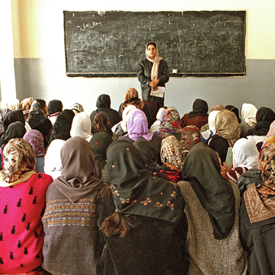 The view from the back of a classroom full of girls wearing hijabs and sitting close together on the floor. The room is plain with just a simple chalkboard on the front wall.