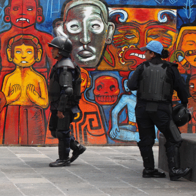 Two police officers in riot gear standing in front of a mural of people in an indigenous Mexican style