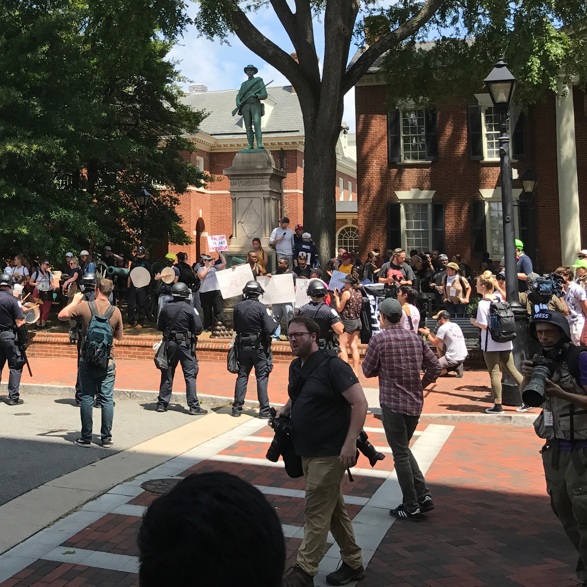 Protestors holding signs in front of a statue of a soldier, surrounded by police.