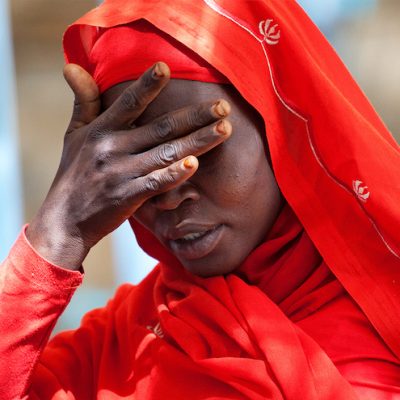 An African woman, dressed in a red dress and head covering, holds her hand over her eyes. She is living in Kassab Camp for Internally Displaced People (IDPs) in Kutum, North Darfur, Sudan, and expressing her sorrow over the increase in rapes in the area to UN representative Ibrahim Gambari in January 2012.